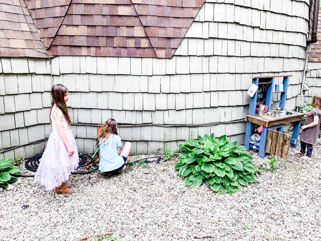rounded exterior cedar wall on geodome home with three little girls playing on pebbles with their outdoor children's mud kitchen